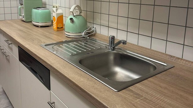 Uma cozinha com pia de aço inoxidável, bancada de madeira, armários brancos e painel de azulejos.