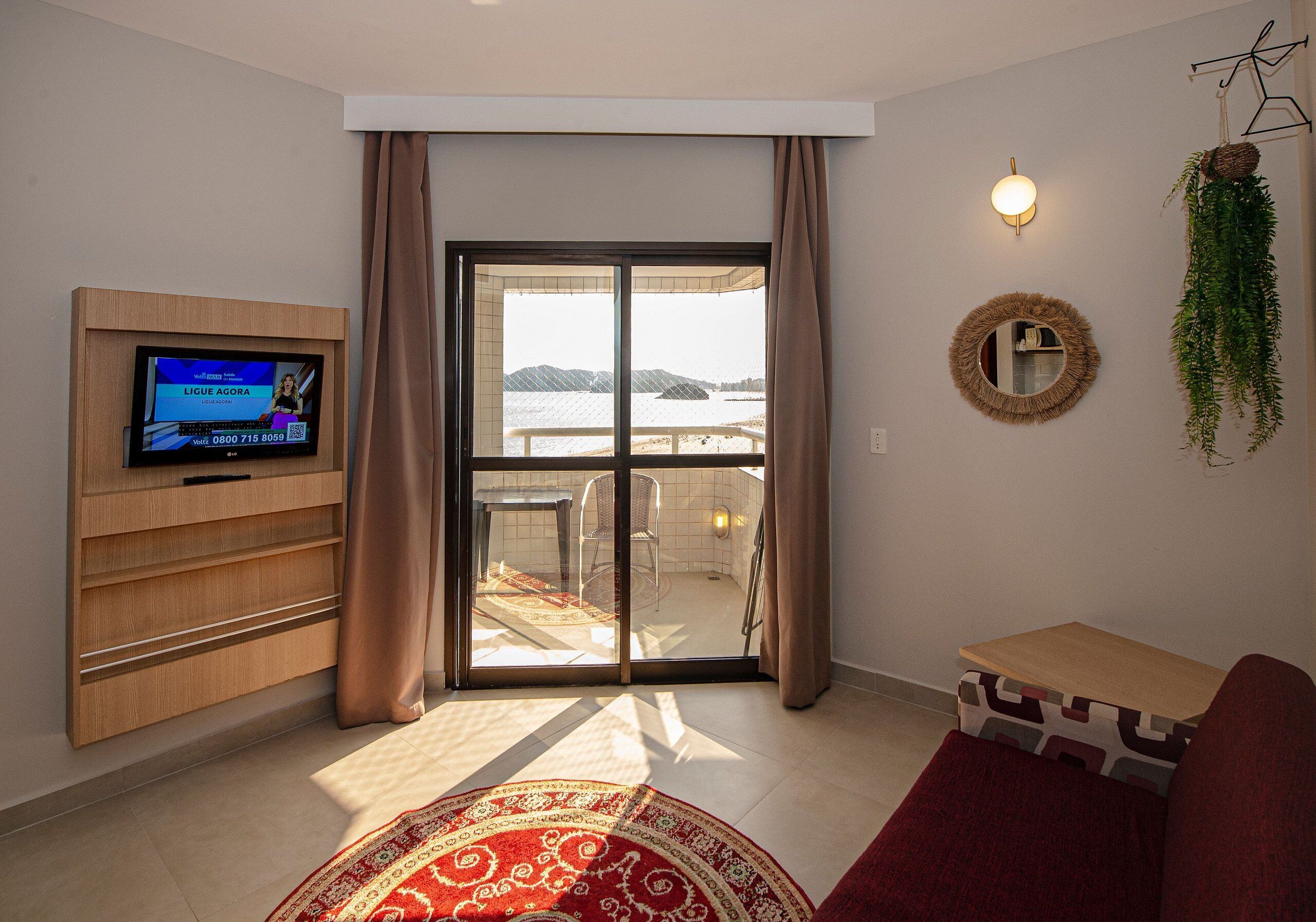 A living room with a television, a sofa, a small table, a sliding glass door to a balcony, and a view of the sea.
