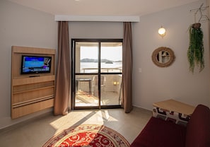 Una sala de estar con televisor, sofá, una mesita, una puerta corrediza de vidrio a un balcón y vista al mar.