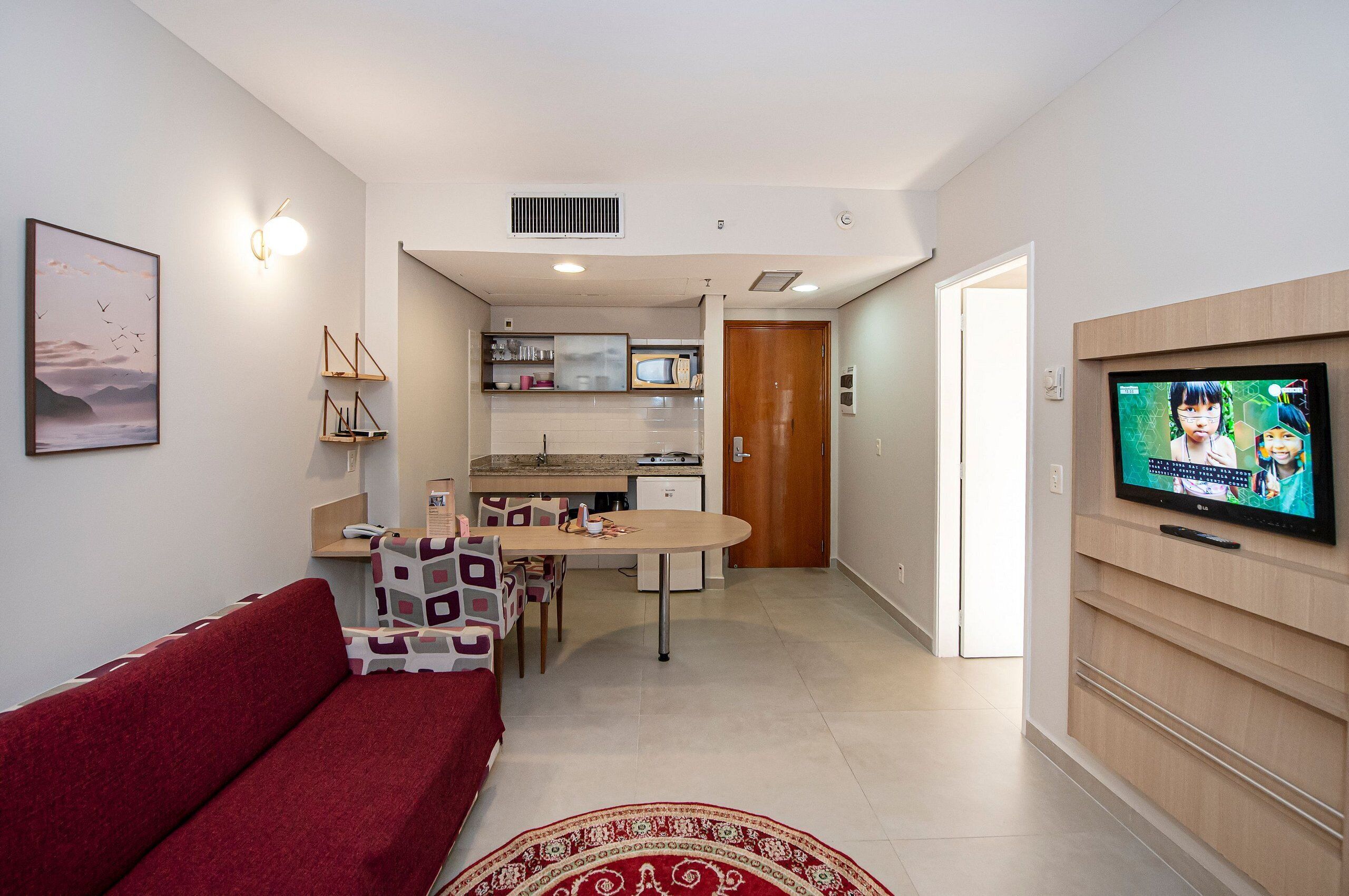 Un espacio habitable compacto con un sofá rojo, una mesa de comedor, una zona de cocina y un televisor montado en la pared.
