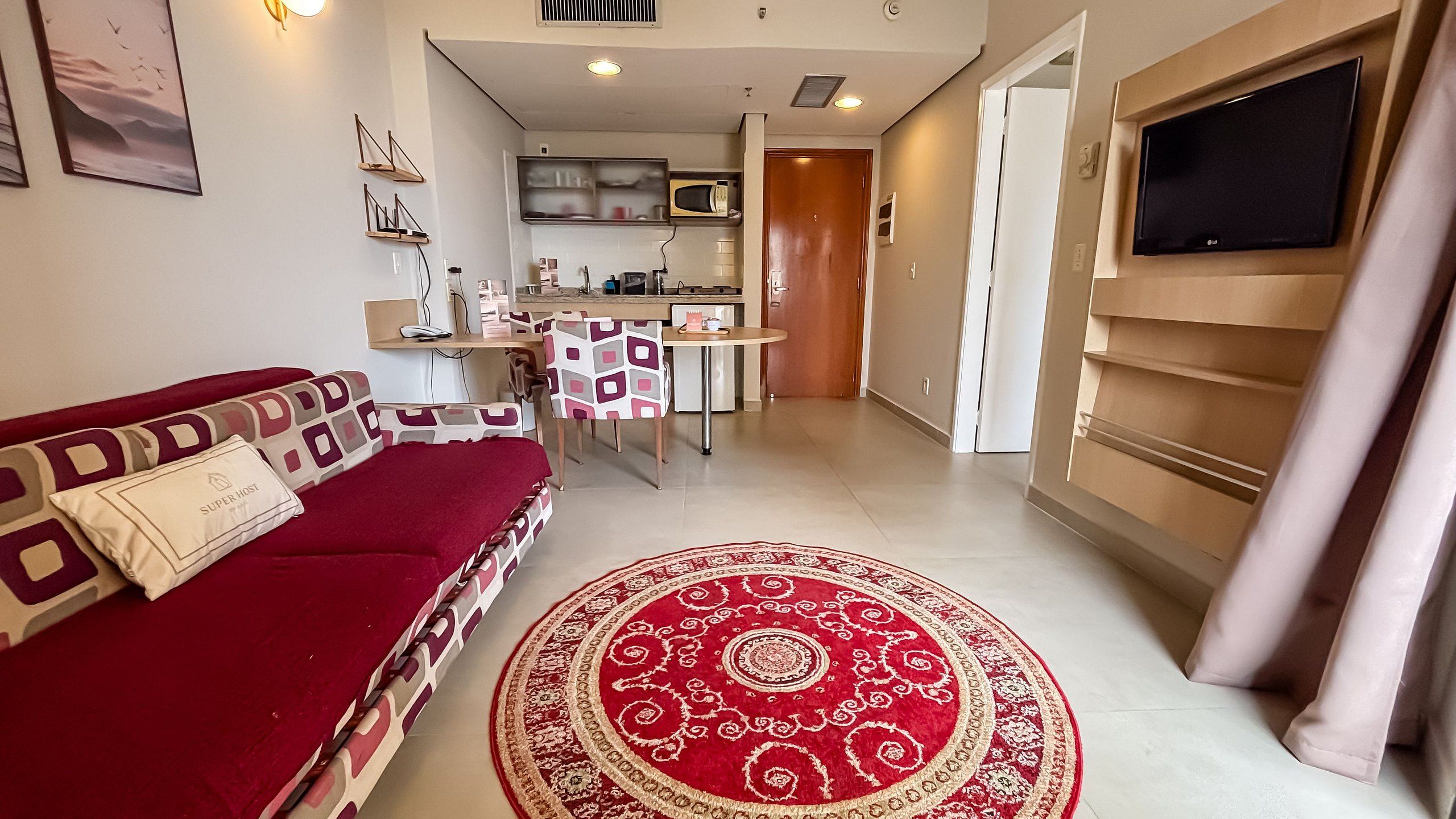 Una sala de estar con un sofá rojo, un tapete estampado, una zona de comedor con mesa y sillas, una cocina con microondas, y un televisor montado en la pared.