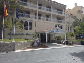 Un hotel de varios pisos con un acceso cubierto, palmeras y una bandera.