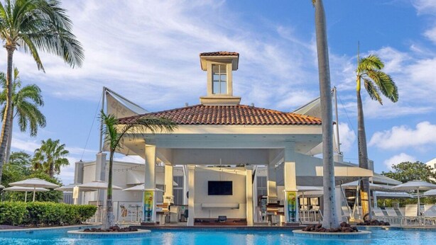 Edifício à beira da piscina com telhado revestido, rodeado por palmeiras e um céu azul límpido.