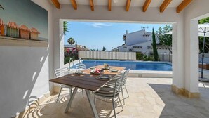 A covered outdoor area with a pool, a dining table with chairs, and a view of a building and a fence in the background. - Boho Villa Montsalvat (Pego)