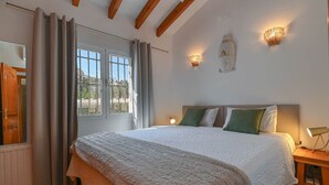 A bedroom with a large bed, a nightstand, a wall-mounted lamp, and a window with curtains. - Boho Villa Montsalvat (Pego)