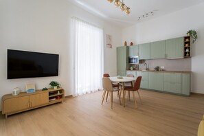 A modern living room with a flat-screen TV, a dining table with chairs, a kitchen with green cabinets, and a large window with sheer curtains. - Santa Luciella Holiday Home  (Naples)