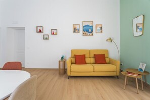 A room with a yellow sofa, a small round table, and a white bed. There are framed pictures on the wall and a lamp on a side table. - Santa Luciella Holiday Home  (Naples)