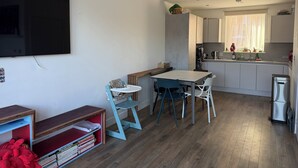 A compact living space with a kitchenette, a dining area with a table and chairs, a television mounted on the wall, and a small bookshelf with books. - Lovely 3-bedroom house in tranquil London perfect for your stay -sleeps 6 (London)