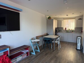 A compact living space with a kitchenette, a dining area with a table and chairs, a television mounted on the wall, and a small bookshelf with books.