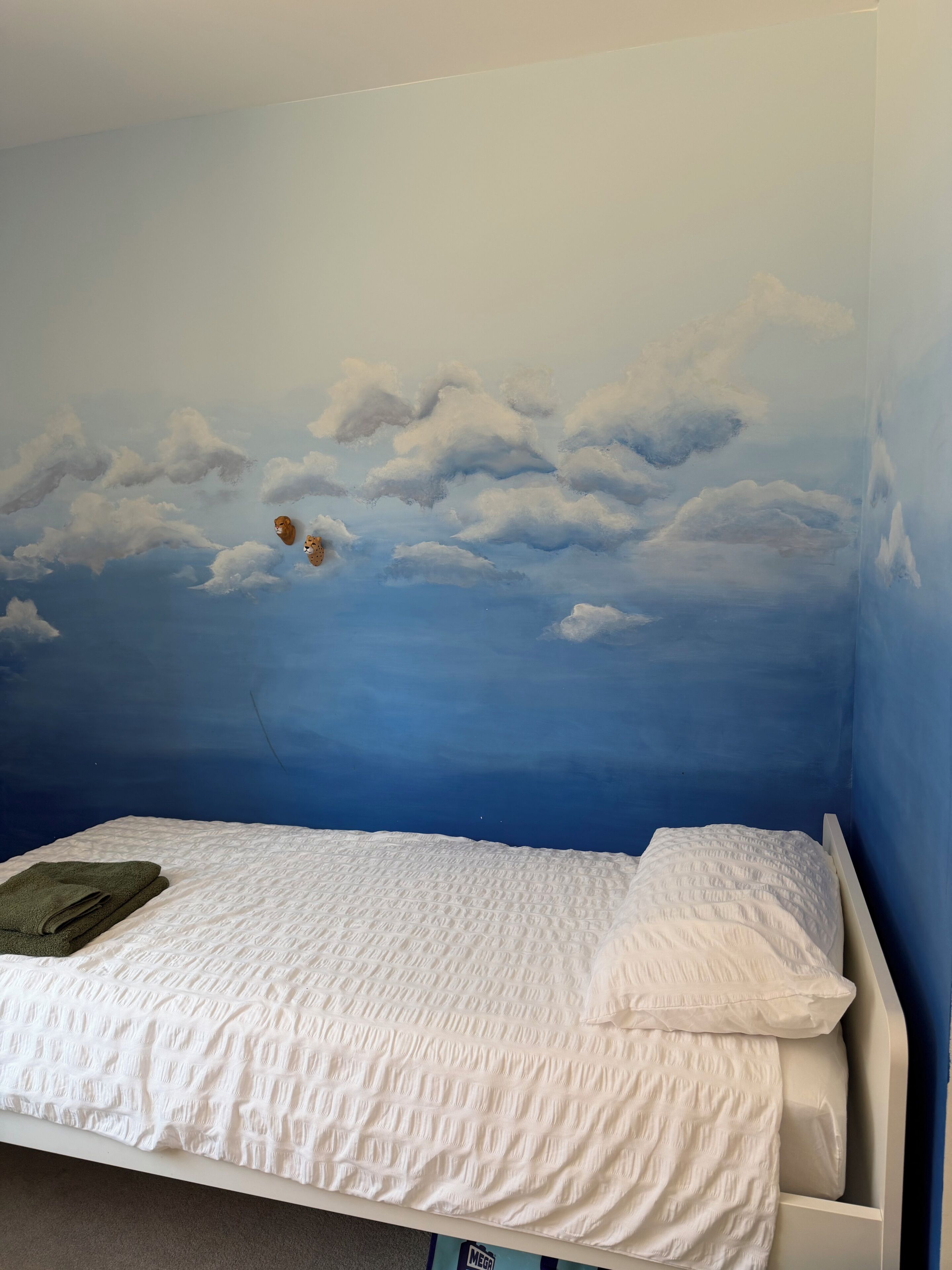 Un dormitorio con una cama, una toalla verde y un mural en la pared con nubes y un avión.