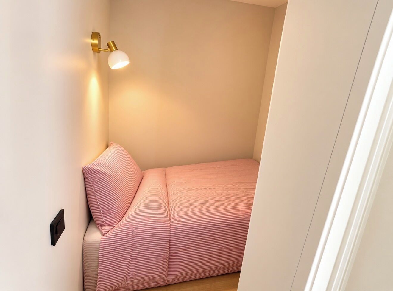 Una habitación pequeña y estrecha con una cama, una lámpara de pared y una puerta.