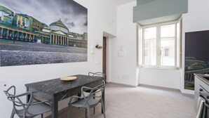 En matplats med ett bord och stolar i marmor, en stor målning som föreställer en stadsbild på väggen och ett fönster med utsikt över byggnader.