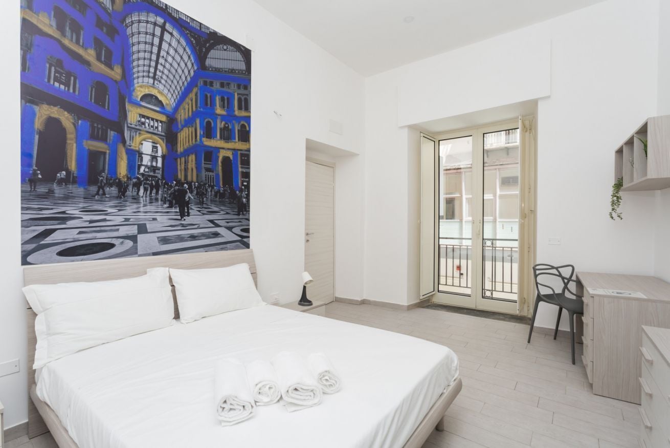 A hotel room with a bed, a desk, a chair, a large window, and a mural on the wall.