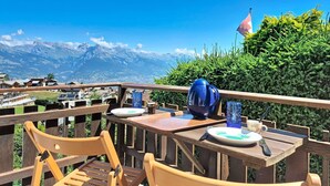 Eine Holzterrasse mit einem Tisch für zwei Personen, mit Blick auf eine gebirgige Landschaft, in der ein Baum eine Flagge trägt.