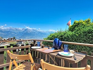 Eine Holzterrasse mit einem Tisch für zwei Personen, mit Blick auf eine gebirgige Landschaft, in der ein Baum eine Flagge trägt.