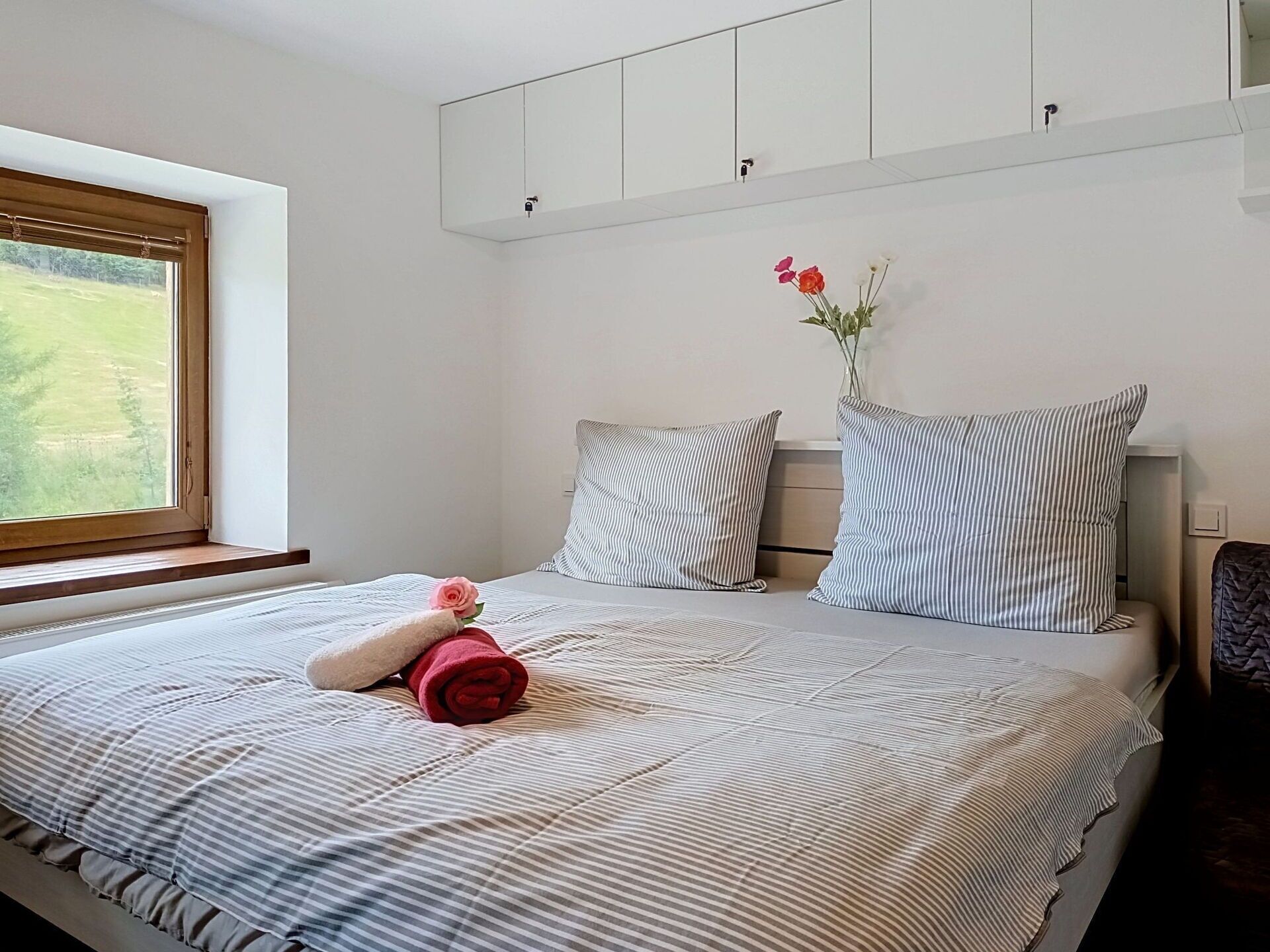 A bedroom with a bed, pillows, a vase with flowers, and a window with a view.