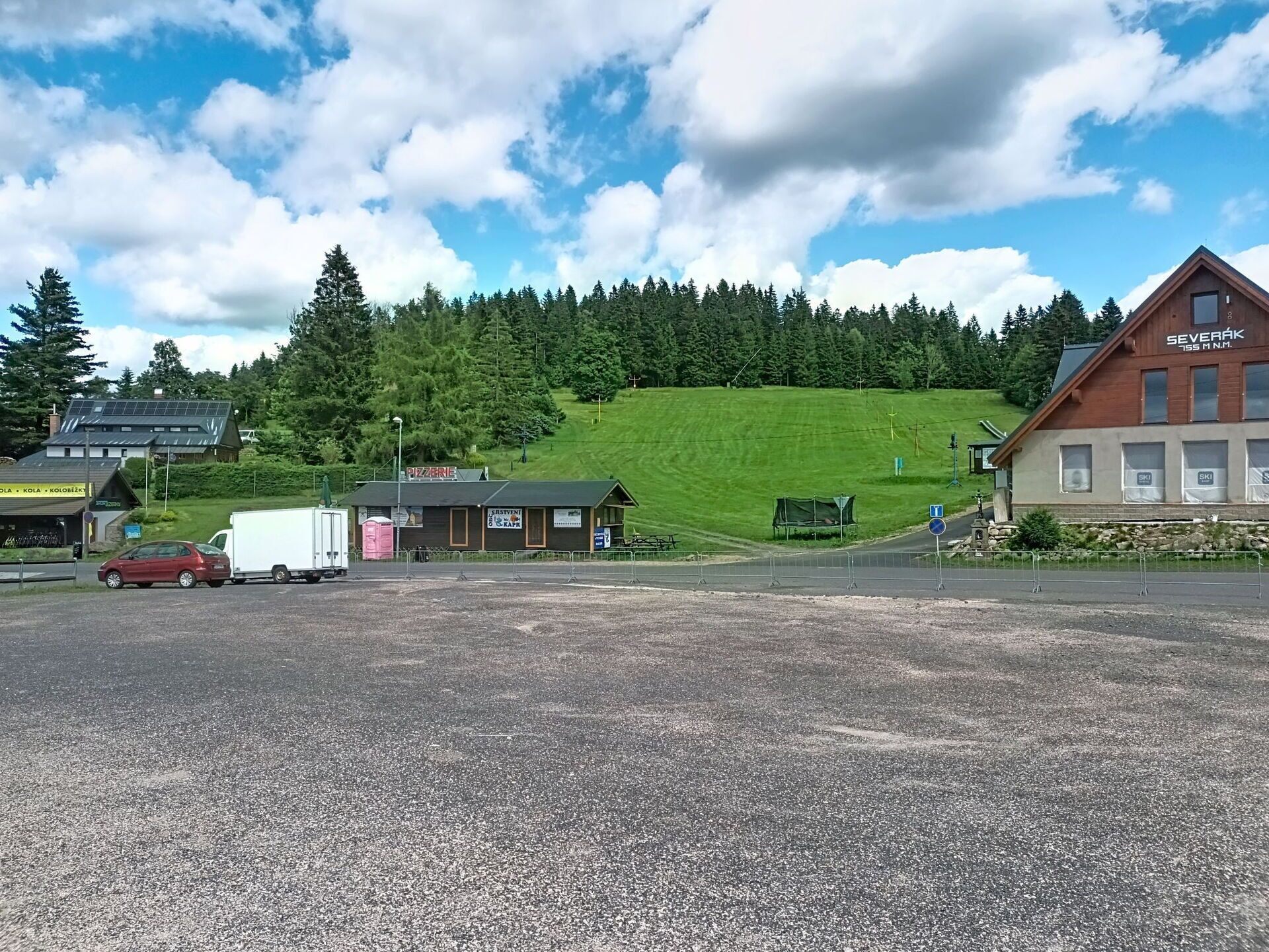 Parkkipaikka, jonka taustalla näkyy rakennus, jossa on kyltti, jossa lukee »SEVERÁK 155 M N.M.». Parkkipaikka on vehreän maiseman ja puiden ympäröimä.