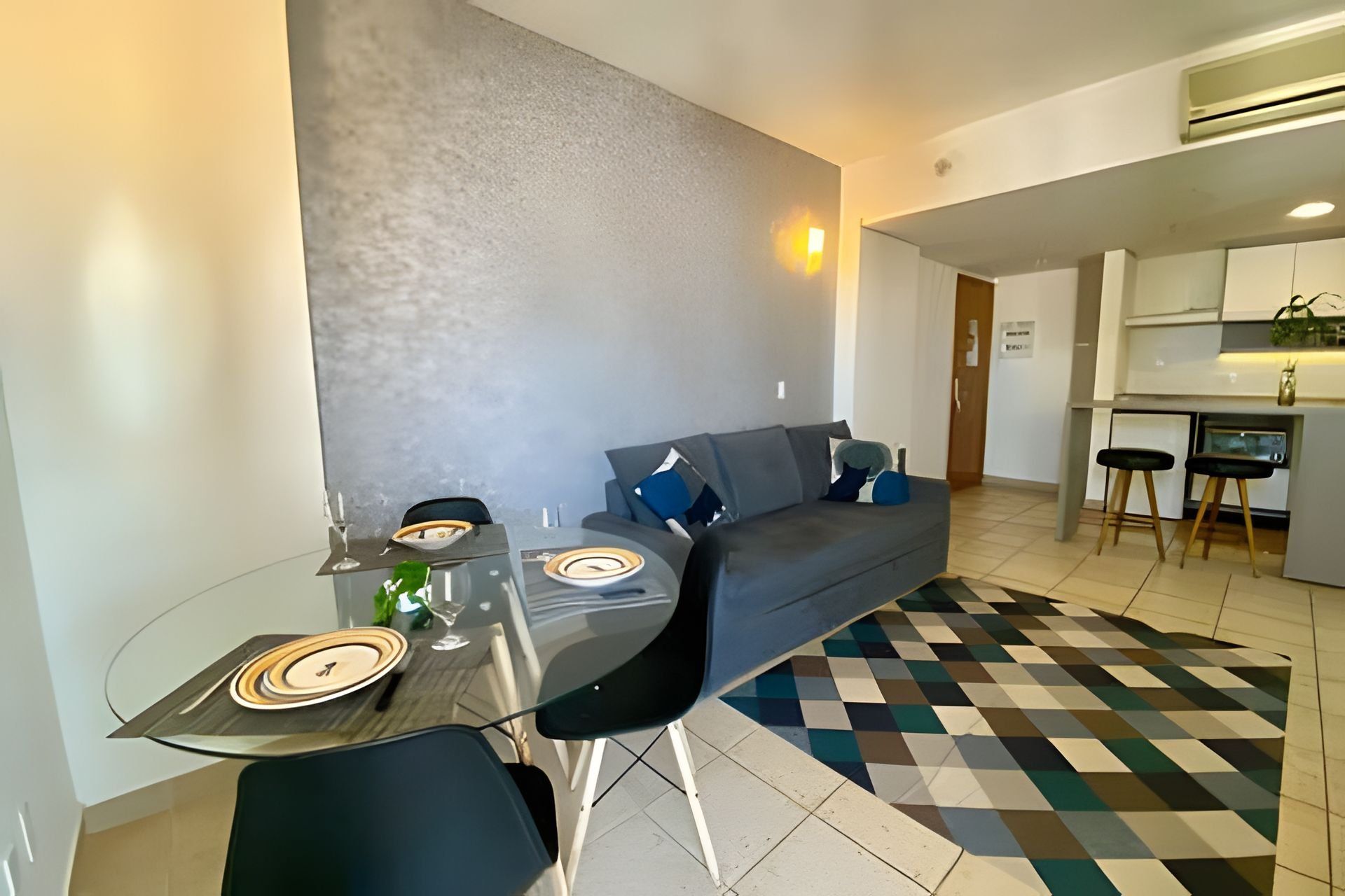Uma sala de estar moderna com uma mesa de jantar de vidro, cadeiras pretas, um sofá cinza com almofadas azuis e um chão xadrez.