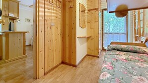 Ein Zimmer mit Holzwänden, einem Bett mit floralem Bettzeug, einem Schreibtisch mit Computer und einem Fenster mit Blick auf Bäume.