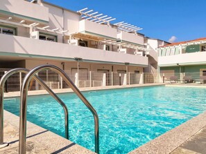 Ein Schwimmbecken mit Metallgeländern, ein überdachter Bereich mit Liegestühlen und ein Gebäude im modernen Architekturstil.