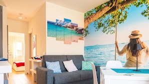 Un saló modern amb un sofà gris, cadires blanques, una taula de dinar i un mural gran amb una escena de platja.