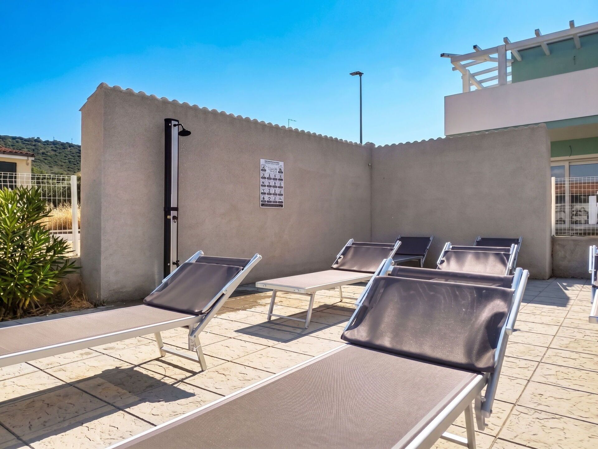 A sunlit outdoor area with several lounge chairs arranged on a tiled floor, a wall with a posted sign, and a building in the background.