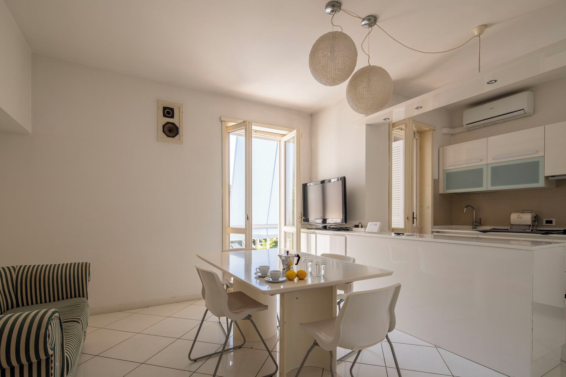 Een moderne keuken met witte kastjes, een zithoek met een witte tafel en stoelen, en een groot raam dat voor natuurlijk licht zorgt.