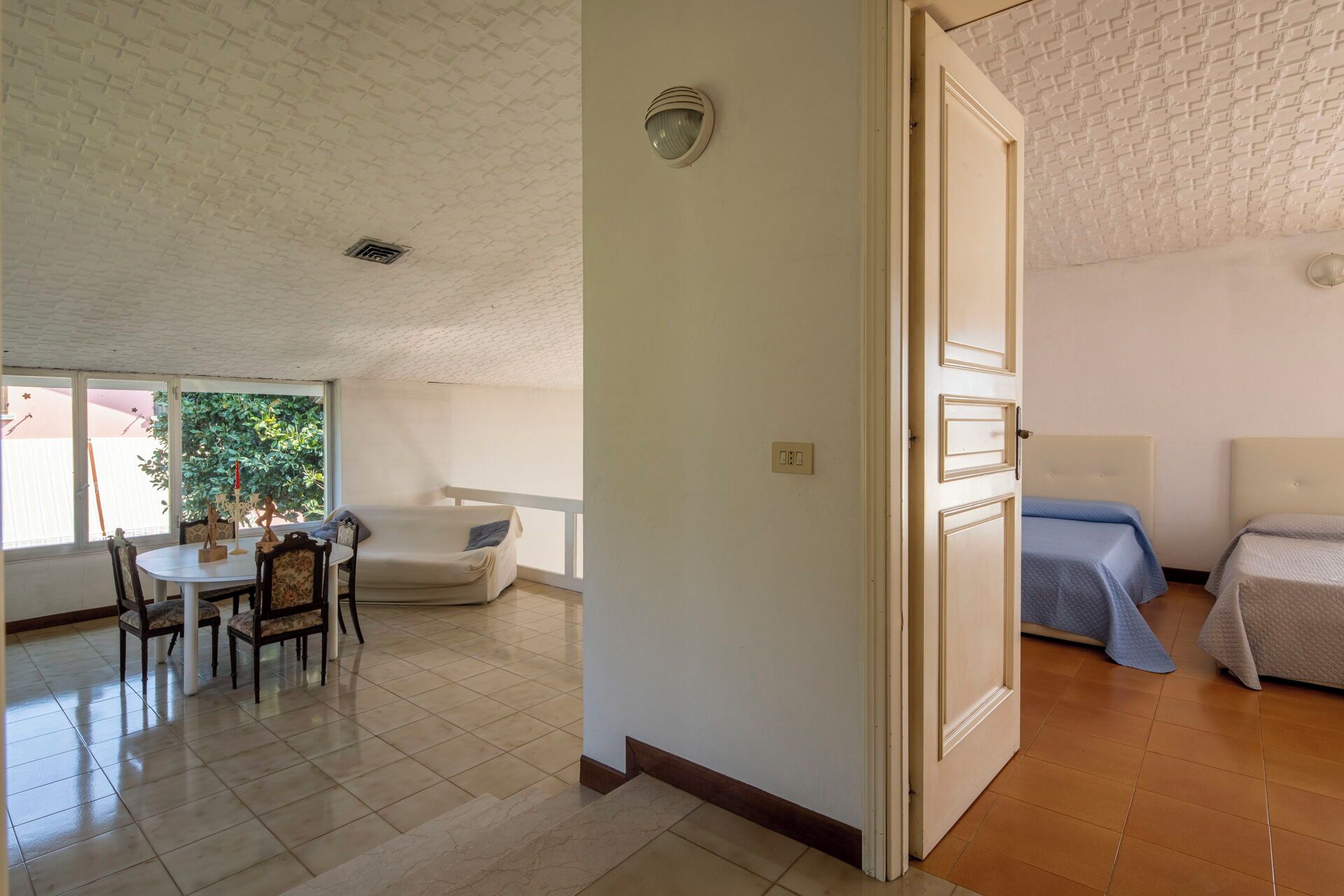 A room with a bed, a dining area with chairs and a table, and a large window with a view of greenery.