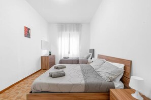 A bedroom with two beds, a wooden floor, a nightstand, a lamp, and a window with curtains. - MA26 [San Siro] Large - New apartment - (Lombardia)