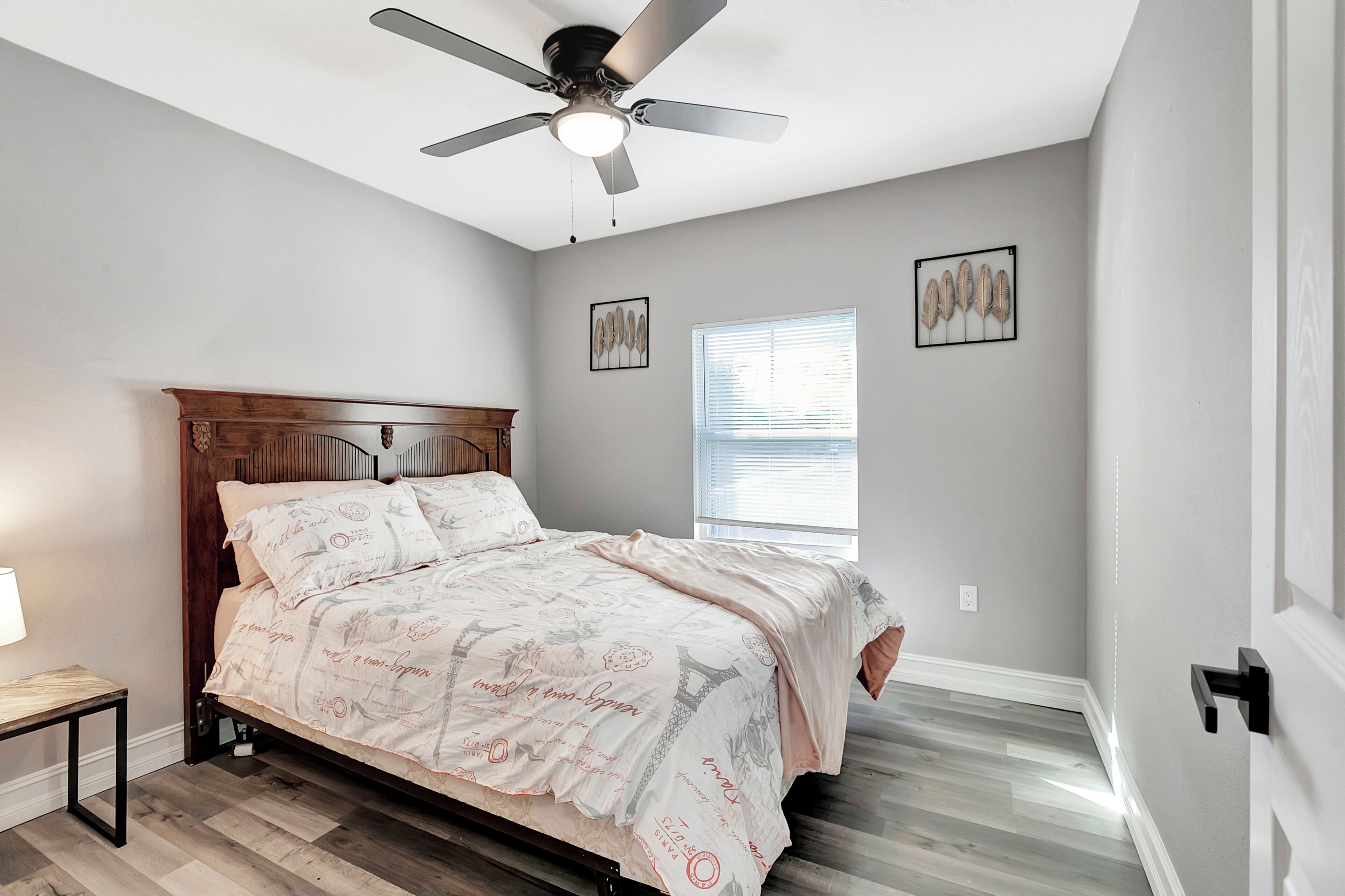 A bedroom with a bed, ceiling fan, window, and artwork on the walls.