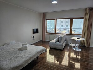 Een hotelkamer met een bed, een bank, een televisie, een raam met gordijnen en uitzicht op gebouwen.