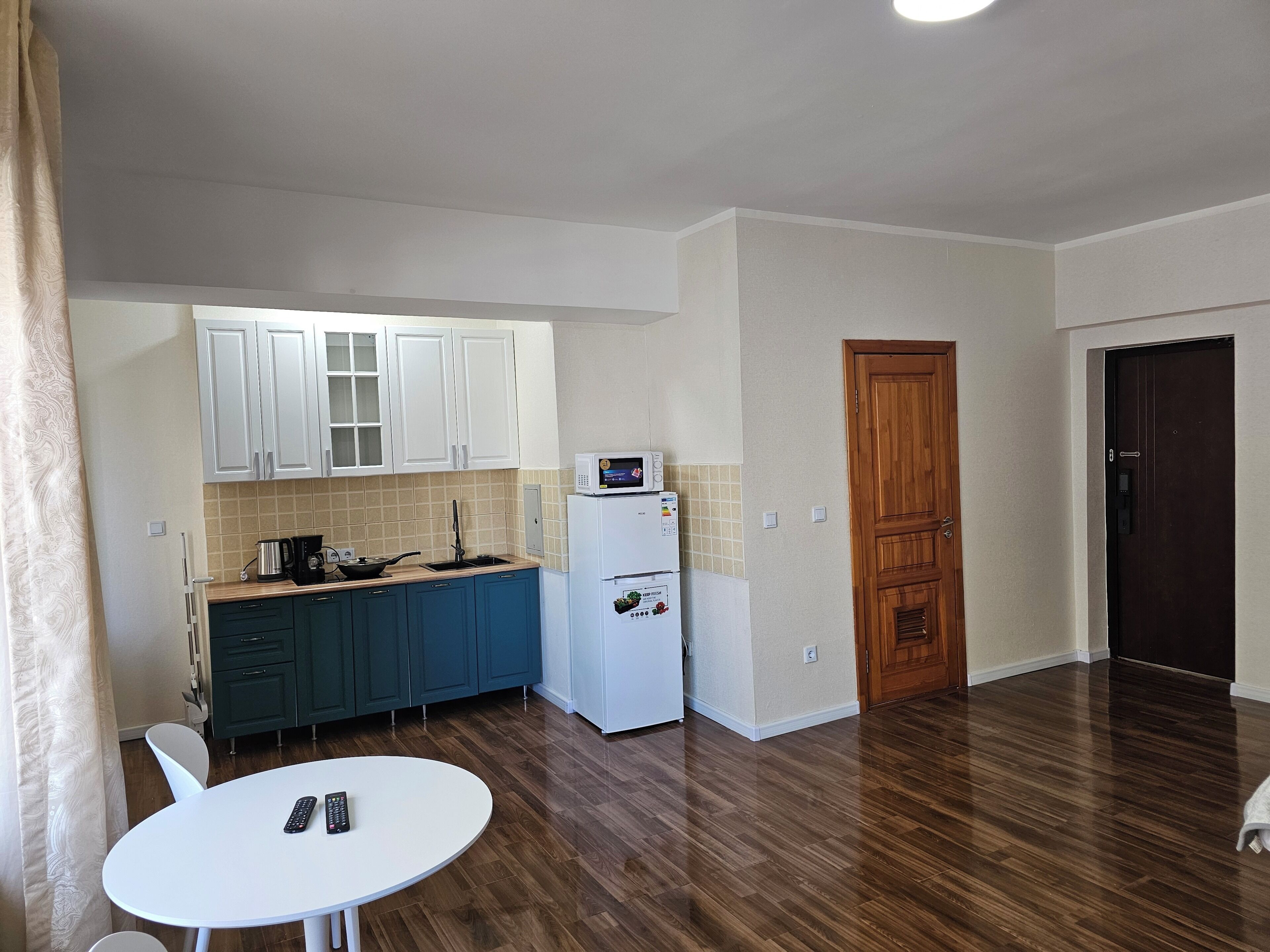 A compact kitchen with white cabinets, a microwave, and a refrigerator. There is a dining table with chairs and a remote control on it.