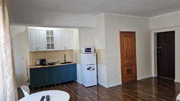 A compact kitchen with white cabinets, a microwave, and a refrigerator. There is a dining table with chairs and a remote control on it.
