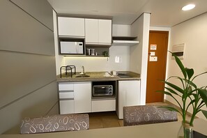 Una kitchenette compacta con armarios blancos, un microondas, un tostador y un horno pequeño. Se observa un lavabo con grifo, y una planta en la esquina.