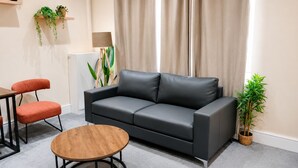 A modern living room with a black leather sofa, a round wooden coffee table, and a red chair. There are two potted plants on shelves and a large window with curtains. - Stylish 3BR House | Free WiFi & Parking | Sleeps 5 | Manchester (Little Hulton)