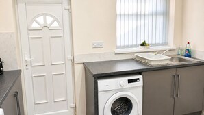 A compact kitchen with a washing machine, a sink, and a window with blinds. - Stylish 3BR House | Free WiFi & Parking | Sleeps 5 | Manchester (Little Hulton)