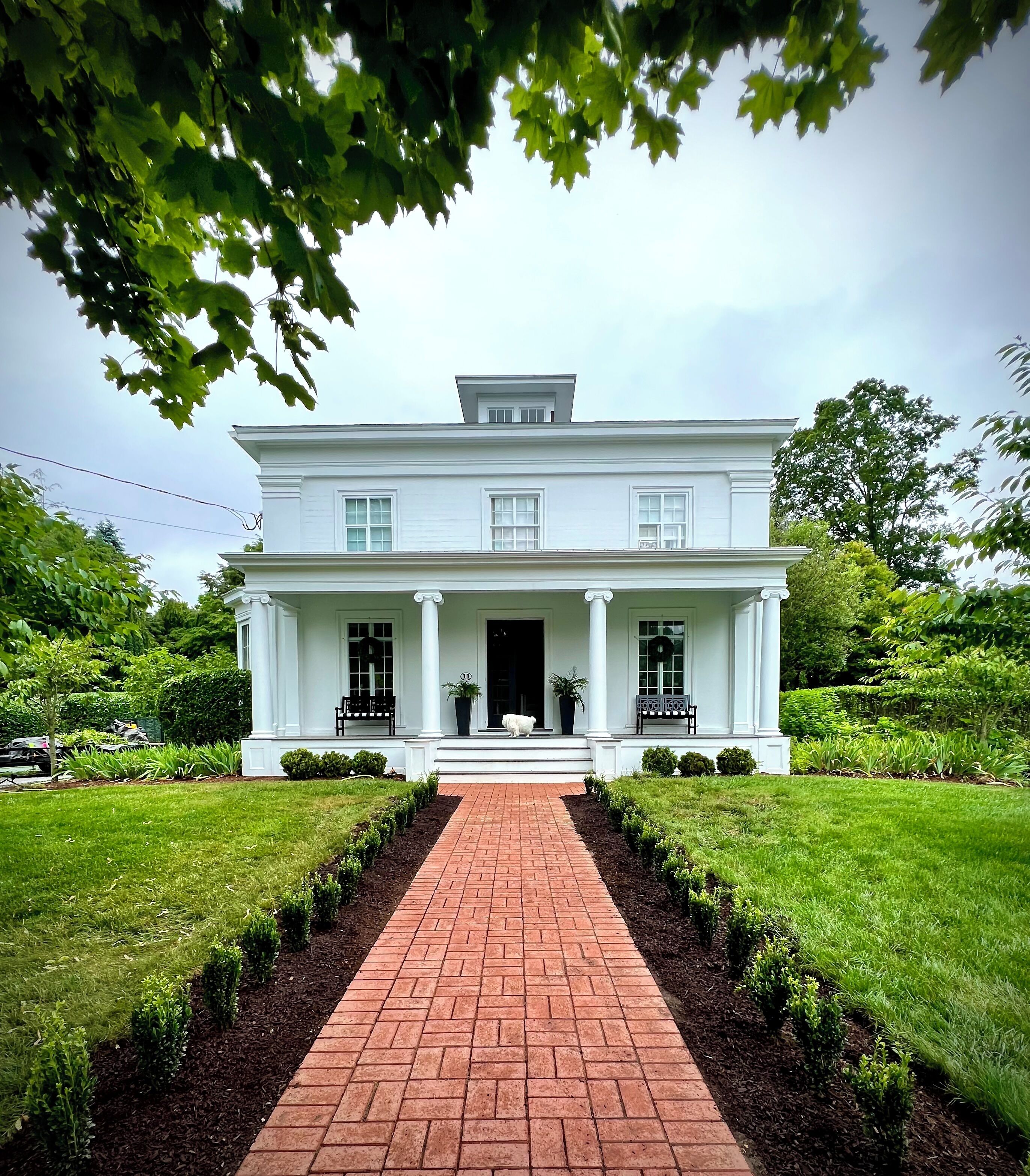 A white colonial-style house with a central front door, two windows on each side, and a brick pathway leading to the entrance.