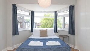 Un dormitorio con cama, dos ventanas, una mesita de noche y una lámpara colgante.