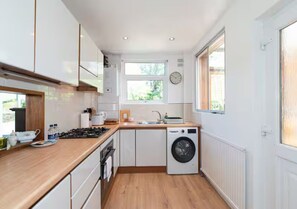 A modern kitchen with white cabinets, a built-in oven, a washing machine, and a window providing natural light. - Charming Home with Garden Near Wembley Stadium - Pass the Keys (London)