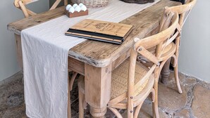 A rustic wooden table with a white tablecloth, surrounded by wooden chairs with woven seats. A basket with a brown bag and a small wooden box are on the table. - Beautiful Cosy 18th Century Cotswold Cottage in Winchcombe (Winchcombe)