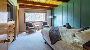 Un dormitorio con techo de madera, una cama grande, dos sillas, una mesita y una ventana con vistas a árboles.