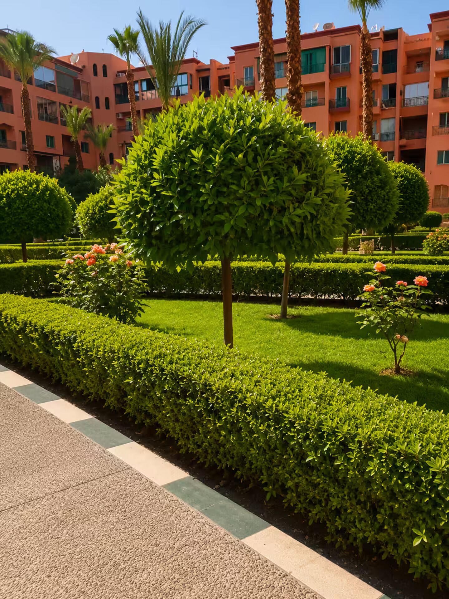 A landscaped garden with neatly trimmed hedges, a variety of trees, and a building with balconies in the background.