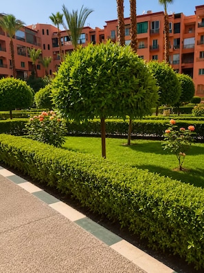 Un jardín arbolado con setos bien cuidados y un edificio con balcones al fondo.