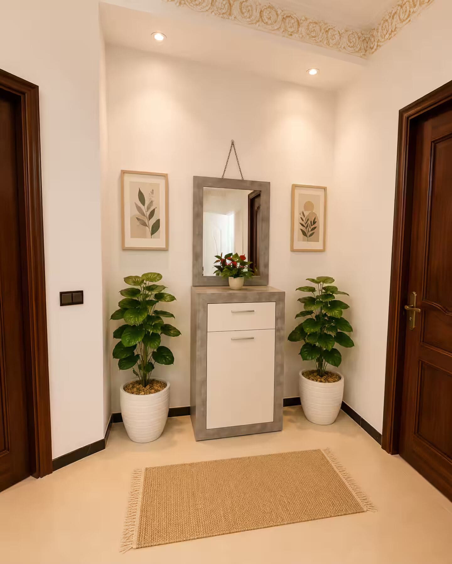 Un couloir avec une petite armoire blanche, deux plantes en pot, des œuvres d’art encadrées accrochées aux murs et un tapis à motifs sur le sol.