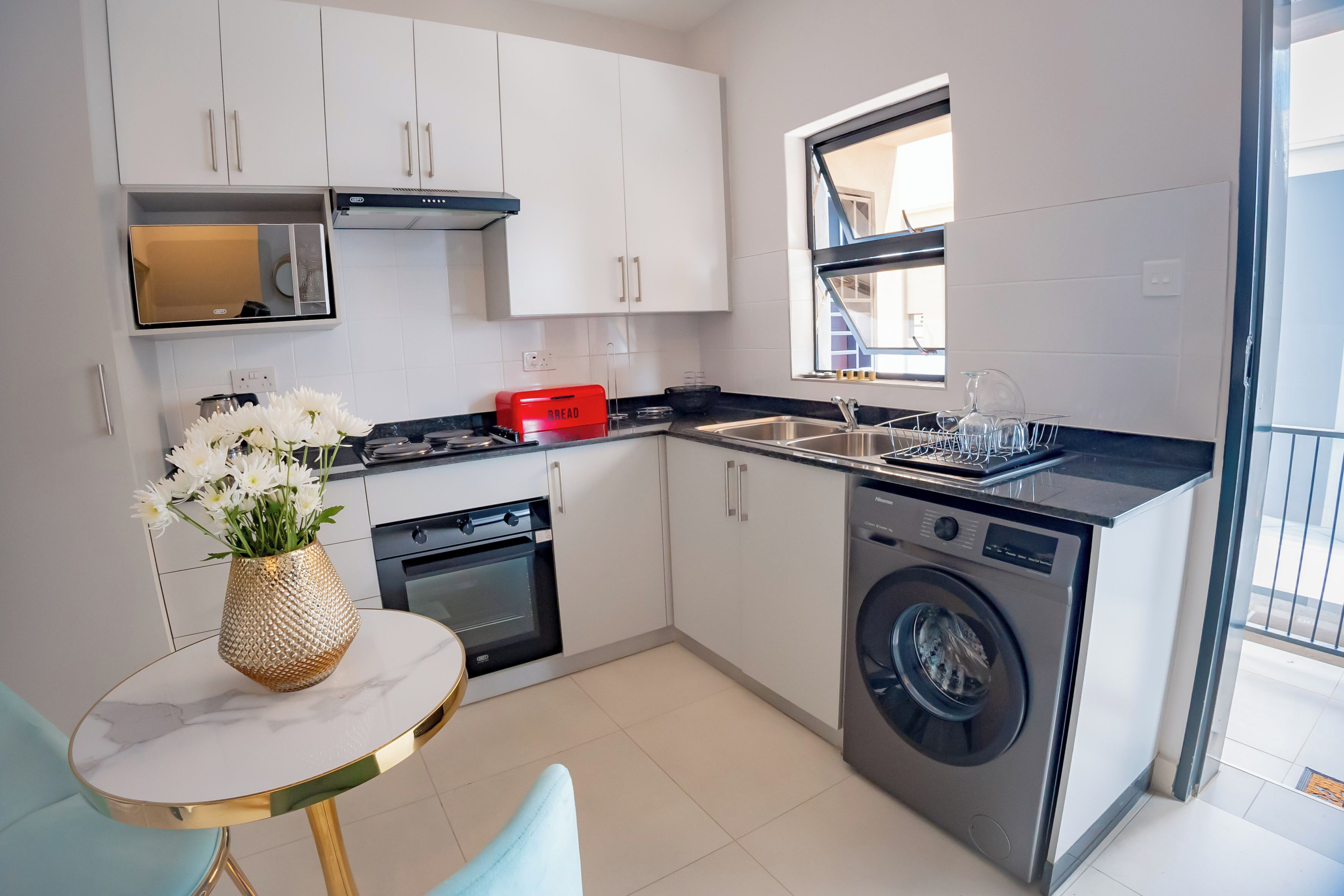 Cocina compacta con alacenas blancas, horno integrado, lavadora y ventana.