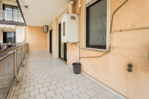 Un couloir avec un sol carrelé, une poubelle et la façade d’un immeuble avec une fenêtre et une porte.