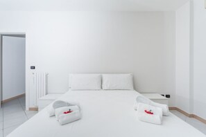 Ein schlicht eingerichtetes Hotelzimmer mit einem großen Bett, weißer Bettwäsche, einem Nachttisch und einer Heizung.