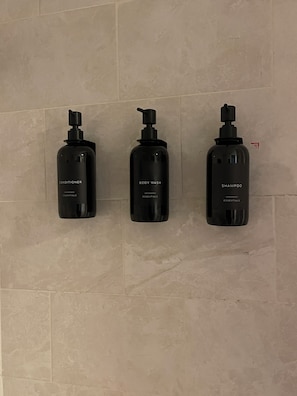 Drei schwarze Flaschen mit Etikett, beschriftet mit Conditioner, Duschgel und Shampoo, auf einer gefliesten Wand.