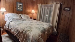 Quarto com painéis de madeira, cama com roupa de cama floral, duas luminárias de cabeceira, mesa de cabeceira e um quadro na parede.
