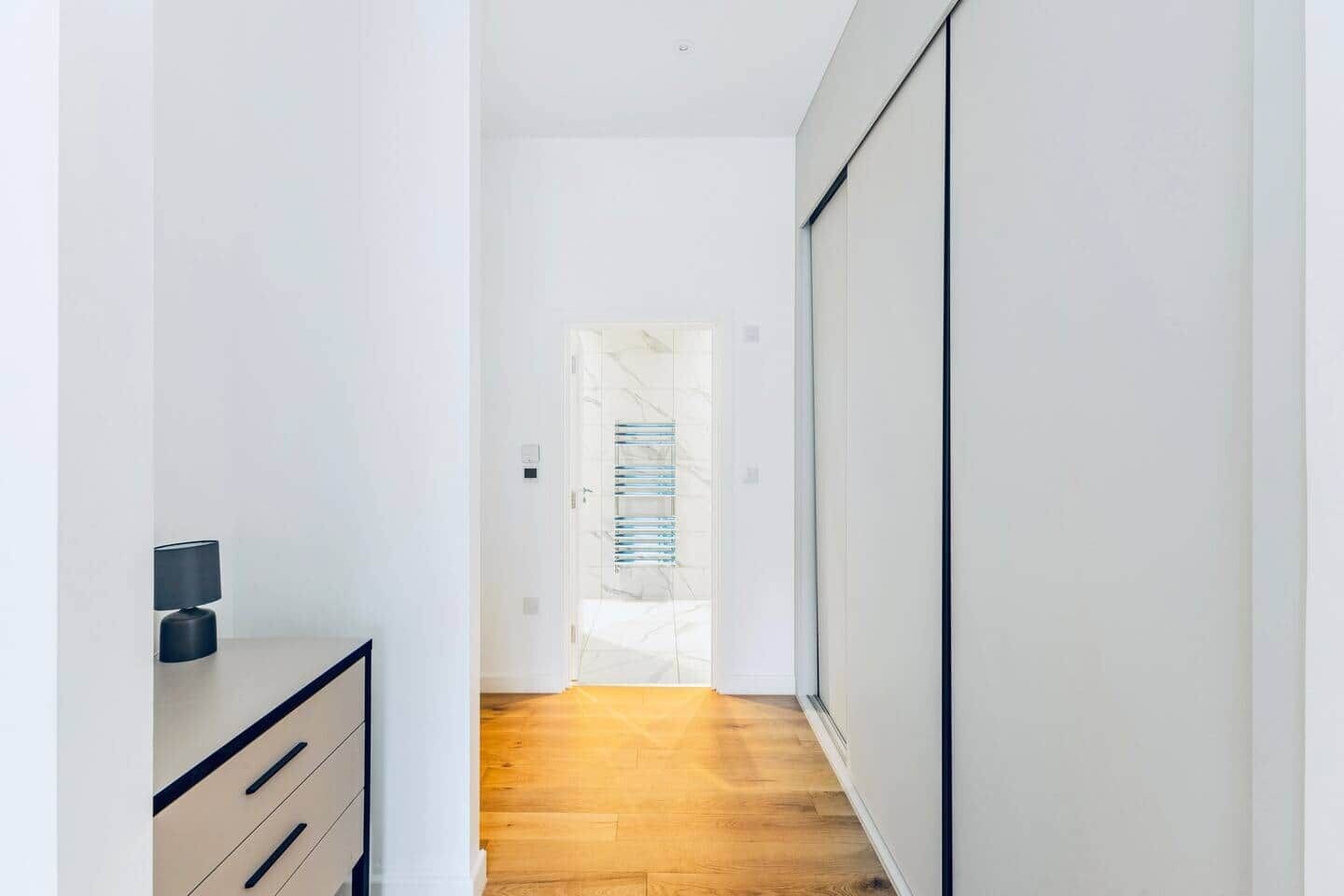 A narrow hallway with a wooden floor, a white wall, and a door leading to a room with a marble wall.